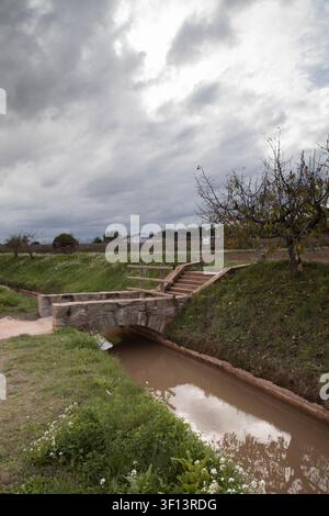 Paesaggio rurale con ponte storico in pietra che attraversa un canale fangoso sotto un cielo nuvoloso Foto Stock