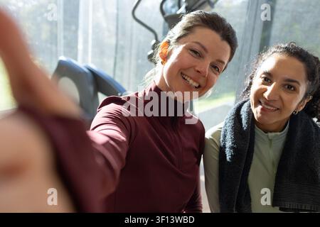 Due amiche sorridenti e in posa per un selfie in palestra, indossano top verde marrone accanto ai tapis roulanti Foto Stock