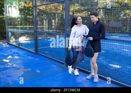 Diverse amiche in abbigliamento sportivo appoggiate a un campo blu con le racchette, che controllano il telefono, spazio di copia Foto Stock