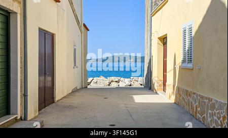 Vista attraverso un vicolo stretto tra edifici beige che si apre su una costa rocciosa, oceano blu e lontane colline verdi sotto un cielo sereno in una giornata di sole Foto Stock