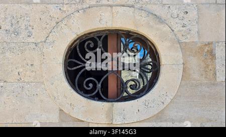 Una finestra circolare inserita in un muro di pietra con una grata in ferro battuto decorativa, che riflette elementi esterni, mostrando dettagli architettonici e rus Foto Stock