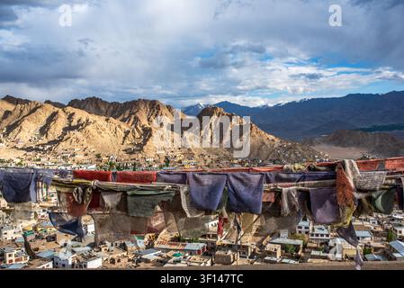Bandiere di preghiera tibetane sfilacciate e colorate sventolano in primo piano, aprendosi su una vasta vista panoramica ad angolazione alta sulla città di Leh e il maestoso Barr Foto Stock