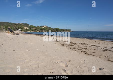 Spiaggia di sabbia idilliaca con canne da pesca piantate nella sabbia e un pescatore su una sedia pieghevole sotto il blu del Mediterraneo, sotto un cielo quasi senza nuvole, Sol Foto Stock