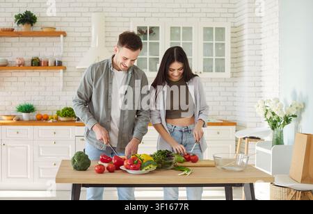 Felice giovane coppia familiare, che cucina insieme l'insalata, si aiuta a vicenda, vegani sani in una cucina moderna a casa Foto Stock