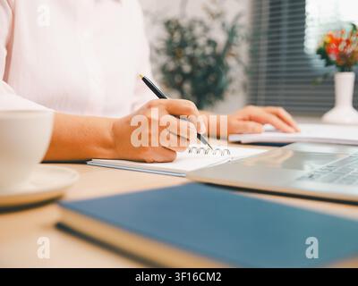 Primo piano della scrittura professionale in un quaderno accanto a un laptop e un mouse sulla scrivania dell'ufficio, che mostra pianificazione, produttività, lavoro remoto, organizzazione aziendale, Foto Stock