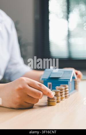 Prima piano di una mano che aggiunge una moneta a monete impilate con un modello di casa blu sullo sfondo, rappresentante la crescita dei risparmi per la casa, il deposito ipotecario e il settore immobiliare Foto Stock