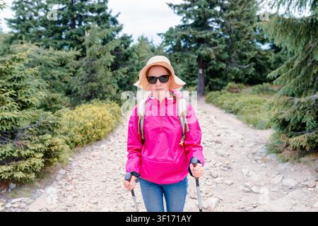 Escursionista in giacca rosa con zaino e bastoni da trekking su un sentiero boschivo roccioso, indossa cappello da sole e occhiali da sole, stile di vita attivo e viaggi all'aperto Foto Stock