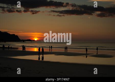 Silhouetti in vista posteriore di persone in piedi su una spiaggia che osservano il tramonto del sole di mezzanotte, Lofoten, Isole Lofoten e Vesterål, Nordland, Norvegia Foto Stock