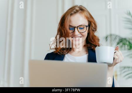 Una donna d'affari dai capelli rossi sorride al computer portatile mentre tiene una tazza di caffè in ufficio Foto Stock