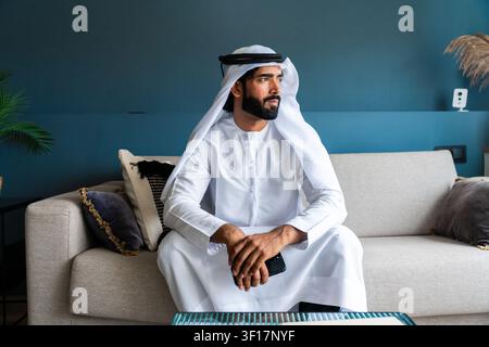 Uomo emirati con barba che indossa ghutra e kandura tradizionali, tiene un telefono, seduto pensieroso su un divano in un soggiorno contemporaneo Foto Stock