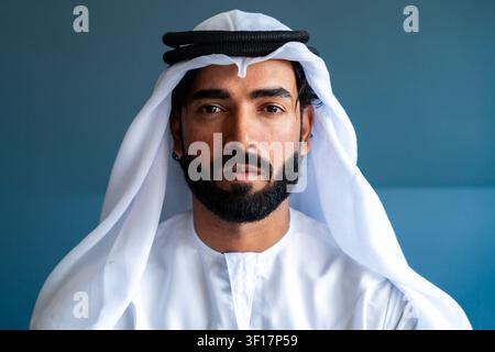 Uomo emiratino in posa con la tradizionale kandura bianca e il ghutra, guarda direttamente nella fotocamera, mostrando un'espressione seria su uno sfondo blu Foto Stock