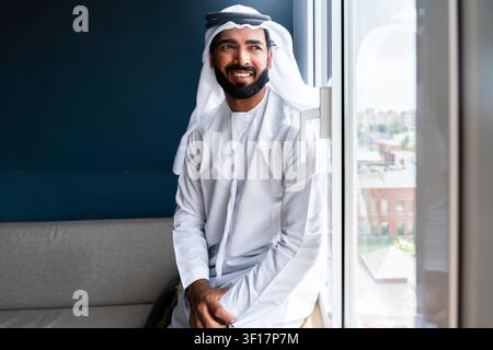 Uomo arabo che indossa una kandura e una ghutra emirati tradizionali, che guarda felicemente fuori da una finestra da uno spazio interno luminoso con una parete scura dietro di lui Foto Stock