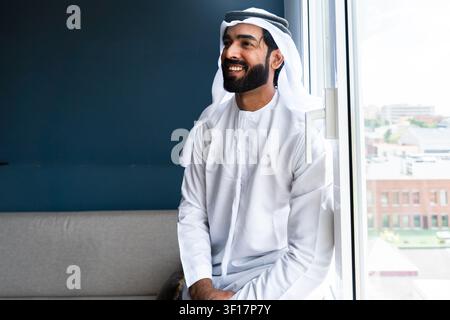 Uomo mediorientale con tradizionale kandura e ghutra accanto a una finestra moderna, guarda lontano pensieroso con un'espressione sicura e felice, riflette Foto Stock
