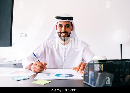 Uomo mediorientale che indossa una kandura e una ghutra emirati tradizionali, sorride direttamente allo spettatore mentre analizza grafici finanziari e prende appunti Foto Stock