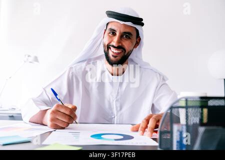 Uomo emirati che indossa una kandura e una ghutra tradizionali sorride alla fotocamera mentre scrive su documenti con grafici e tabelle, rappresentando il successo negli affari Foto Stock