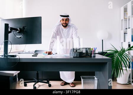 Uomo professionista emirati con tradizionale kandura e ghutra, in piedi alla sua scrivania moderna, si sente sicuro e guarda la fotocamera Foto Stock