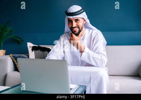Uomo d'affari arabo che indossa una kandura e una ghutra emiratina tradizionali, sorride mentre digita su un laptop, lavorando o studiando dal suo ambiente moderno e confortevole Foto Stock