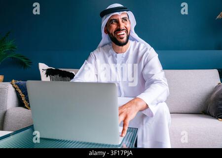 Uomo emiratino che indossa la tradizionale kandura e ghutra, sorridendo allegramente mentre lavora su un laptop da un ambiente domestico confortevole, impegnato in Remo Foto Stock
