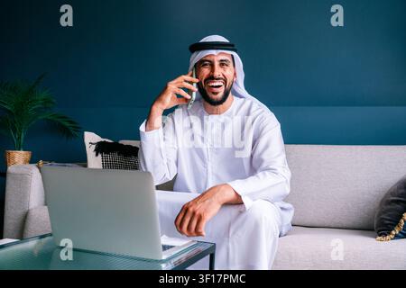Uomo emirati che indossa kandura e ghutra ride mentre parla al telefono, seduto su un divano con un laptop sul tavolino, lavora da un mod Foto Stock