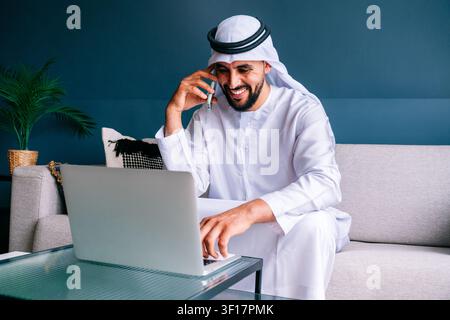 Uomo emiratino che indossa kandura e ghutra, felicemente impegnato nel lavoro a distanza, parlando al telefono cellulare e digitando su un laptop dal suo soggiorno moderno Foto Stock