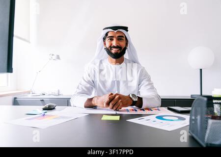 Uomo arabo sicuro di sé che indossa una tradizionale kandura e ghutra, si sente di successo mentre è seduto alla sua scrivania con documenti e grafici in un ufficio contemporaneo Foto Stock