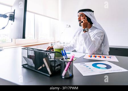 Uomo d'affari arabo che indossa una tradizionale kandura emiratina seduto alla sua scrivania. Sta analizzando rapporti sulle vendite e grafici delle spese mentre parla al cellulare Foto Stock
