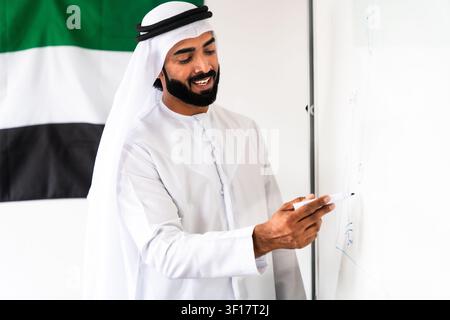 Uomo emirati che indossa una kandura e una ghutra e sorride. Presenta idee e scrive su una lavagna bianca. Con la bandiera degli Emirati Arabi Uniti sullo sfondo Foto Stock