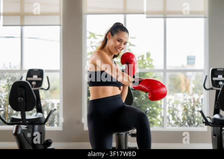Giovane donna sicura di sé con guantoni da boxe rossi che si diverte con l'allenamento di boxe e il lavoro di piedi in palestra. Scena per coaching personale, viaggio di perdita di peso, quotidiano w Foto Stock