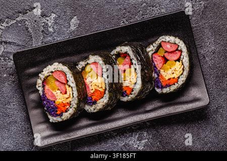 Un piatto nero pieno di kimbap (o gimbap) con ripieni colorati. Gli ingredienti includono salsiccia, cetrioli, uovo e verdure. Foto Stock