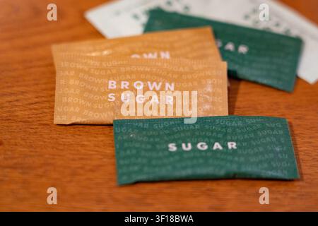 Sacchi di zucchero individuali in carta verde e marrone etichettati "Zucchero" e "Zucchero di canna", sparsi ordinatamente su una superficie calda di legno di un tavolo da caffè Foto Stock