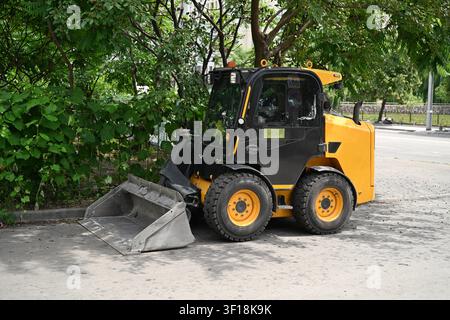 Caricatore frontale giallo parcheggiato su una strada asfaltata vicino ad alberi verdi per la manutenzione urbana. Foto Stock