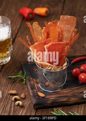 Fette e strisce di carne affumicata essiccata in un secchio di metallo con un bicchiere di birra, noci e spezie su un tavolo di legno Foto Stock