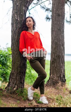 Donna sicura di sé in camicetta rossa e pantaloni olivastro si appoggia a un albero in un parco verde, ritratto casuale a figura intera con luce naturale Foto Stock