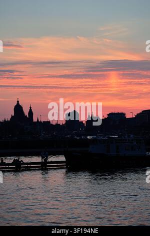 Edifici silouettati sul lungofiume di Amsterdam e una linea di tetti a cupola si ergono sotto un cielo vivacemente striato di arancione e rosa, riflessi in acque scure e increspate. Foto Stock