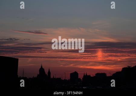 Una bassa linea dell'orizzonte di Amsterdam forma una silhouette nera sotto nuvole striate arancioni e rosse del tramonto, con una banda orizzontale di luce pallida sopra l'acqua increspata. Foto Stock