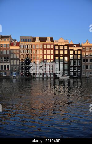 Vista verticale delle alte e strette case del canale Damrak ad Amsterdam, le cui facciate calde si riflettono nell'acqua blu scuro del canale sotto un cielo azzurro senza nuvole. Foto Stock