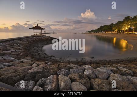 Vista serena del mattino da un molo di pietra su una baia tranquilla a Sanur Beach all'alba, con le luci dei resort che si riflettono nell'acqua immobile a Bali Foto Stock