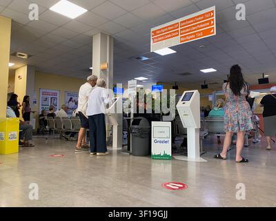 Cremona, Italia, 19 giugno 2026 Clienti del settore sanitario che navigano in una sala d'attesa moderna, utilizzano chioschi self service e consultano segnaletica informativa in un pu Foto Stock