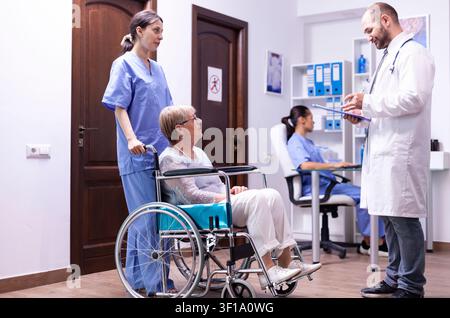 Medico che ha una conversazione con una donna anziana disabile Medico che controlla la diagnosi di una donna anziana disabile in sedia a rotelle. Personale medico nel corridoio dell'ospedale con una paziente disabile. Foto Stock