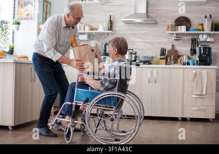 Uomo anziano che aiuta la moglie a svuotare la borsa della spesa, persone mature con verdure fresche dal mercato Vivere con una persona disabile con problemi di deambulazione. Foto Stock