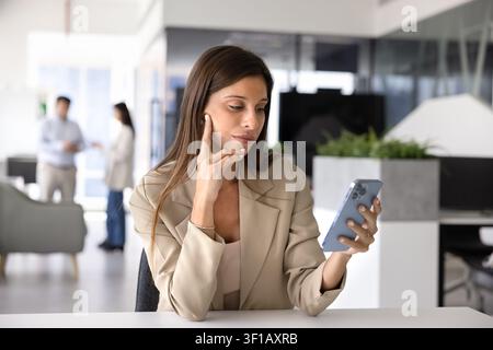 Donna latina pensierosa seduta in un ufficio aziendale con smartphone Foto Stock