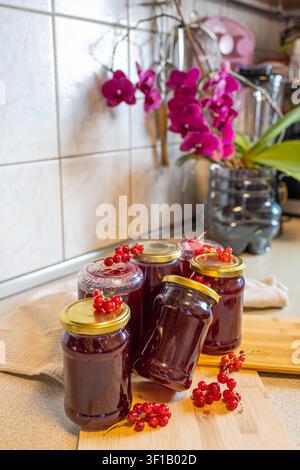 Marmellata fatta in casa di ribes rosso in barattoli di vetro con bacche fresche e orchidee in fiore in una cucina rustica, conserve tradizionali di frutta estiva Foto Stock