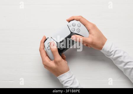 Persona che tiene un controller bianco moderno per videogiochi con entrambe le mani, vista dall'alto su uno sfondo di legno bianco, il dispositivo elettronico presenta vari ma Foto Stock