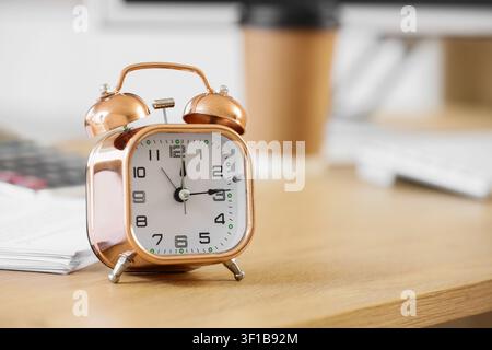 Sveglia in rame su una scrivania di legno con una tazza di caffè sfocata e documenti sullo sfondo, l'orologio ha un quadrante bianco con numeri neri chiari Foto Stock