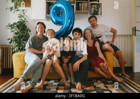 Famiglia multigenerazionale felice riunita su un divano durante la festa di compleanno di un ragazzo a casa, con un grande palloncino blu a forma di nove e un cane domestico. Foto Stock