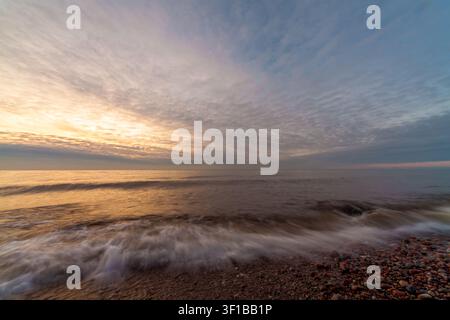 Mare calmo all'alba con luce dorata soffusa, nuvole strutturate e onde gentili che si infrangono su una riva di ciottoli. Paesaggio costiero tranquillo con ampio orizzonte Foto Stock