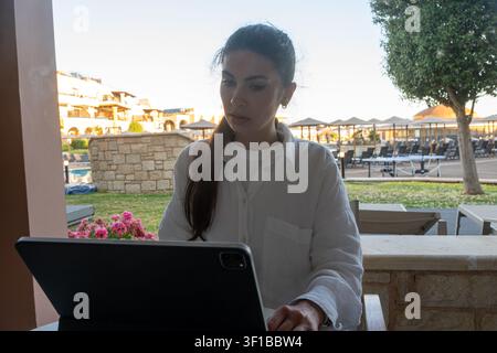 Giovane donna bruna in una camicia di lino bianca si siede su una terrazza all'aperto di un resort e lavora su un tablet durante il giorno. Foto Stock