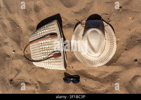 Vista aerea degli accessori da spiaggia estivi tra cui una borsa cappello in paglia intrecciata e occhiali da sole neri disposti su una spiaggia sabbiosa alla luce del sole. Foto Stock