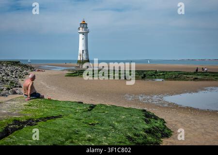 Un uomo è seduto su una roccia muschiosa, godendosi la giornata soleggiata vicino alla piscina sabbiosa di New Brighton. Il faro si erge in alto sullo sfondo, con onde gentili Foto Stock