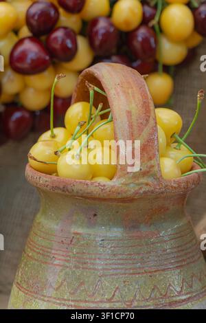 Ciliegie gialle dolci disposte in una giara di terracotta rustica con ciliegie miste sullo sfondo, creando una natura morta di frutta estiva tradizionale con atmosfera rurale Foto Stock